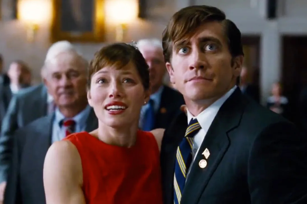 Jessica Biel in a sleeveless dress and Jake Gyllenhaal in a suit and tie stand together in a crowded room, both looking up with seemingly surprised expressions