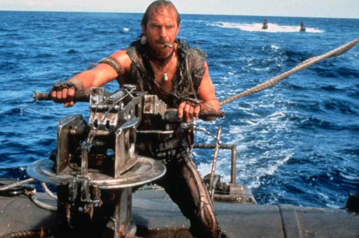 A man is standing on a boat in the ocean, operating a heavy piece of machinery, with a cigar in his mouth. Two boats are visible in the background