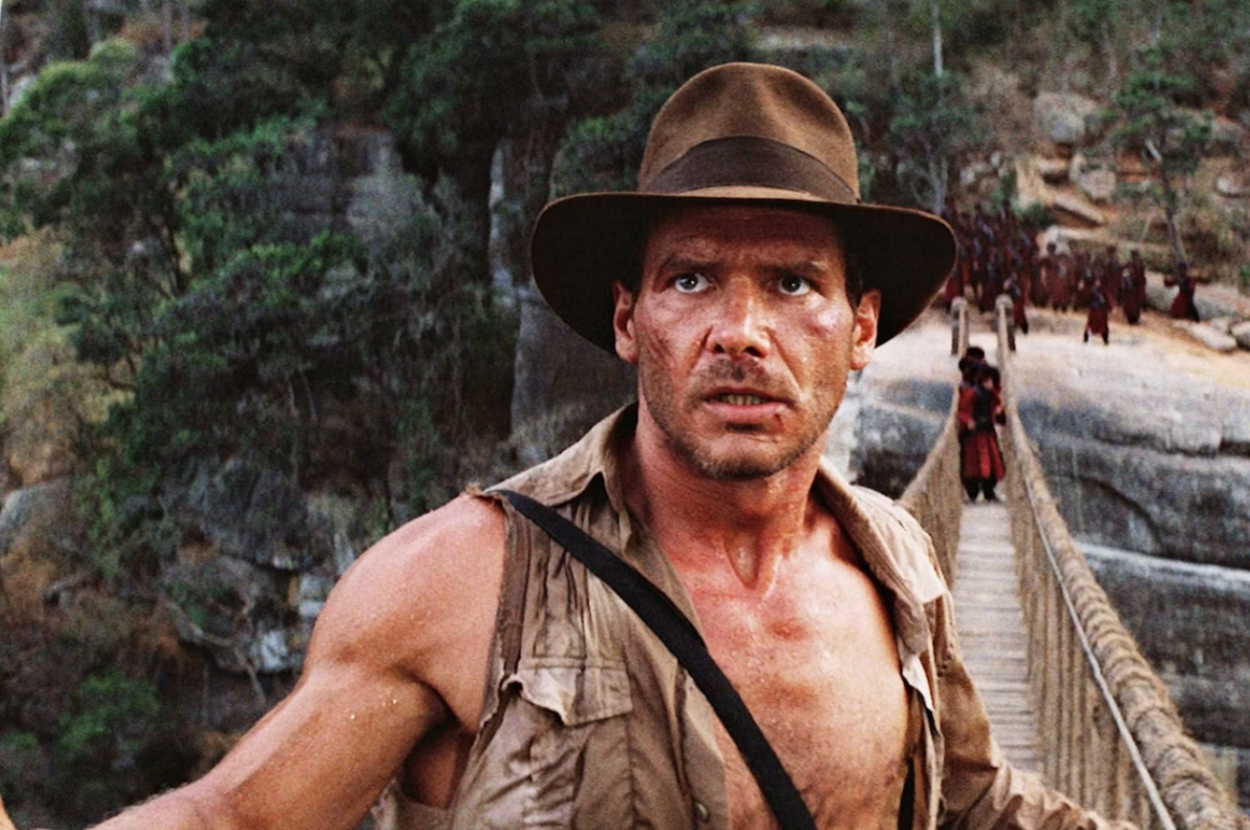 Harrison Ford, dressed as Indiana Jones in a rugged sleeveless shirt and hat, looks concerned while standing on a rope bridge in a jungle setting