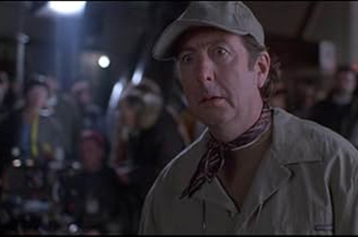 A man in a light jacket and cap, with a scarf tied around his neck, looks surprised at something off-camera while a crowd in the background is blurred