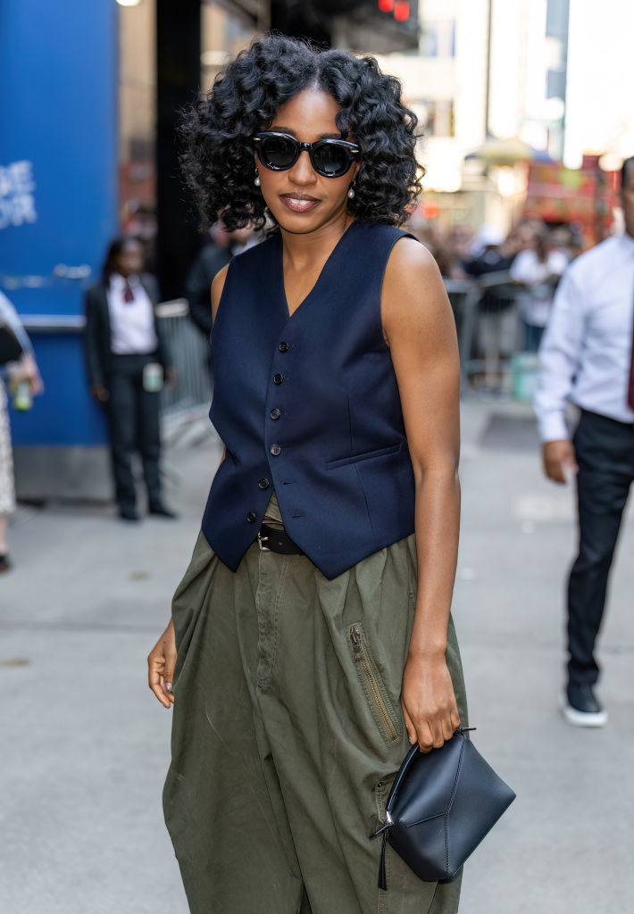 A person with curly hair, wearing sunglasses, a sleeveless vest, and loose-fitting pants, holds a black clutch while walking outdoors