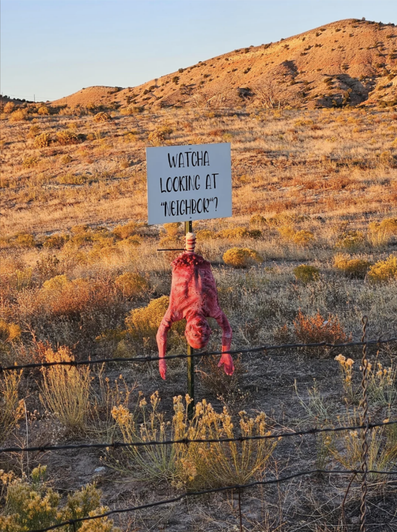 A sign in a desert-bearing landscape reads, “Watcha looking at, neighbor?” with a fake bloody torso hanging below