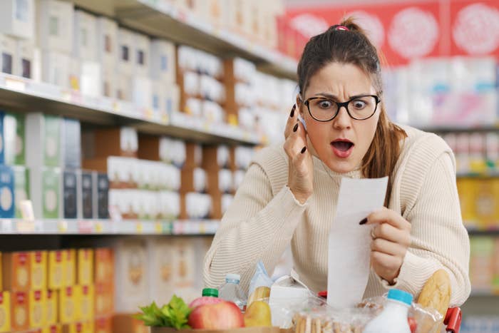 A shocked woman in a grocery store holding a receipt while pushing a shopping cart filled with various items