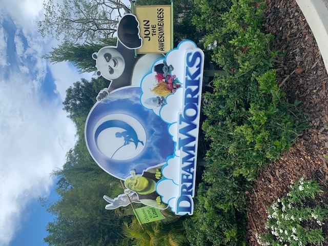 Sign with DreamWorks characters (Shrek, Po the Panda, Donkey, Puss in Boots) and text, "Join the Awesomeness," set in a garden with lush greenery