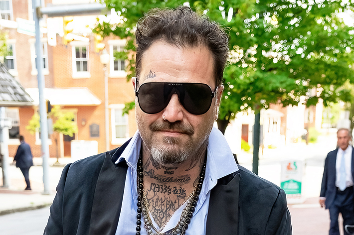 A man with tattoos and a beard, wearing dark sunglasses, a black jacket, a light shirt, and a beaded necklace, walks on a street with buildings in the background