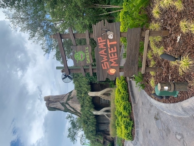 Image shows a wooden sign reading "Shrek's Swamp Meet" with painted characters from Shrek. In the background, there is a cottage with a tree-like appearance