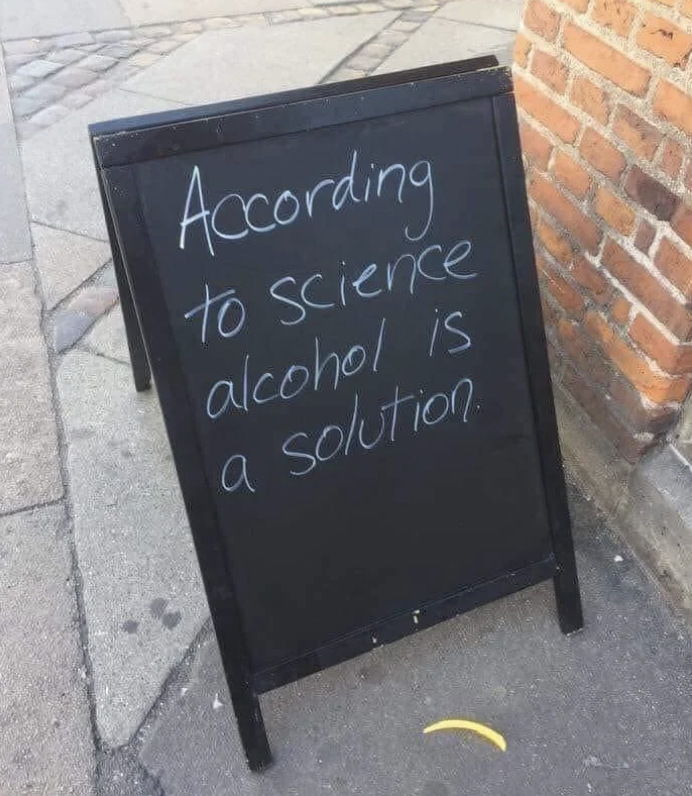 A black sidewalk sign reads, &quot;According to science alcohol is a solution.&quot; The sign is placed outside on a paved street