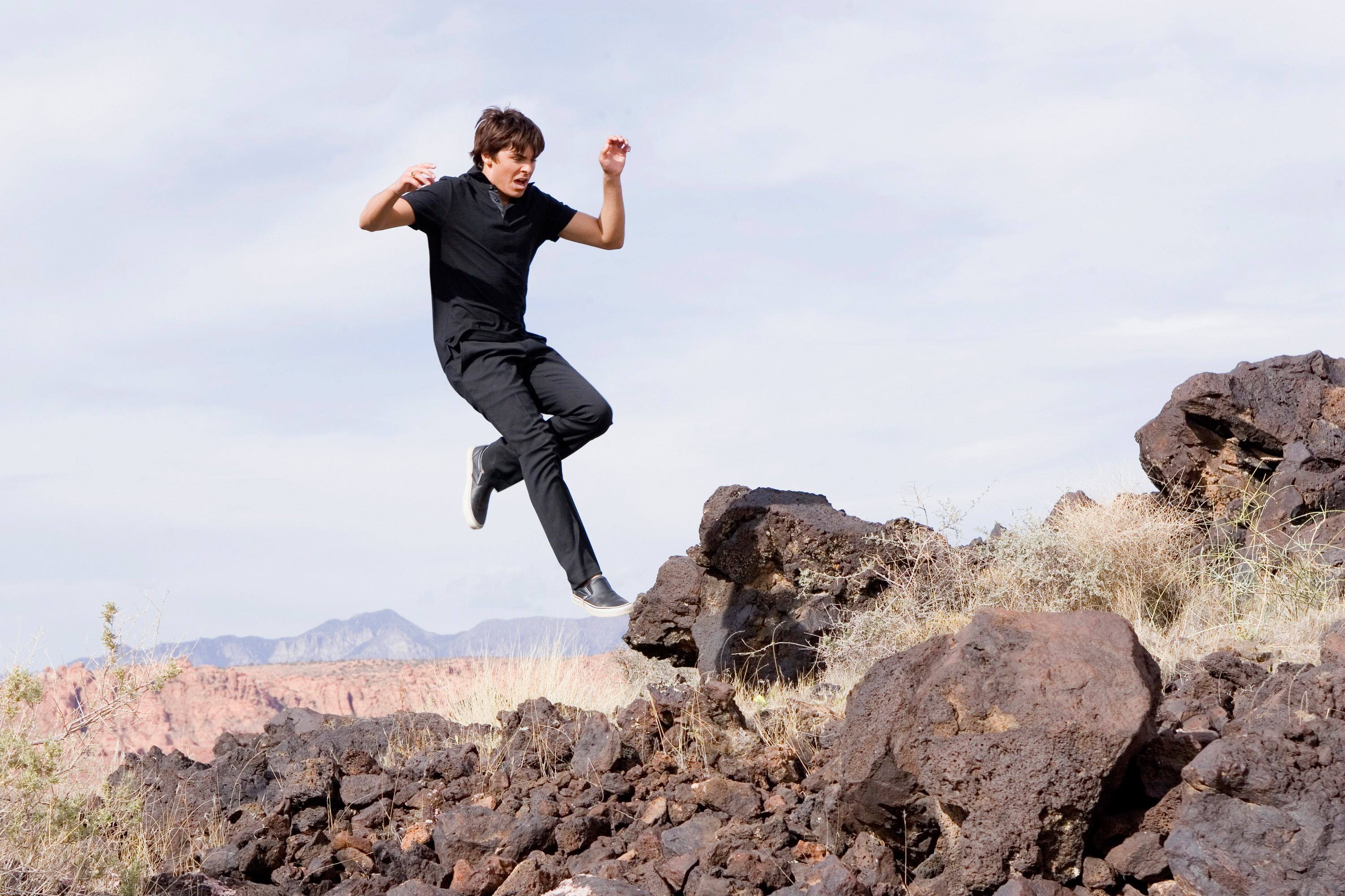 A person dressed in a black T-shirt and black pants is captured mid-air while jumping over rocky terrain in an outdoor setting