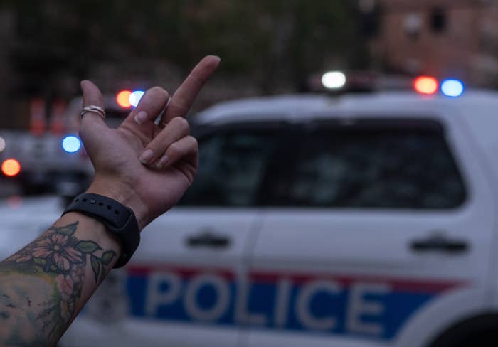A tattooed arm in the foreground flips off a distant police car with flashing lights
