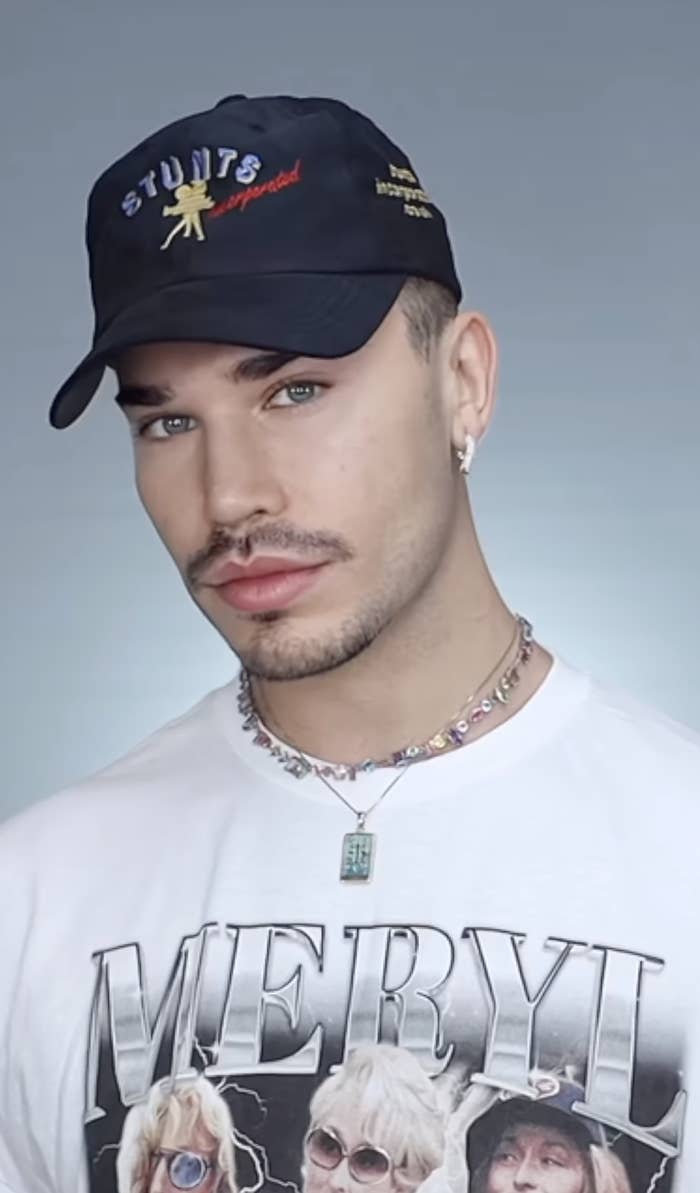 A person wearing a &quot;Stunts inspired&quot; cap and a graphic T-shirt with &quot;MERYL&quot; text and images. They have silver earrings and a layered necklace