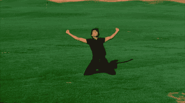 A person in all black clothing celebrates on their knees on a grassy field with arms raised in victory