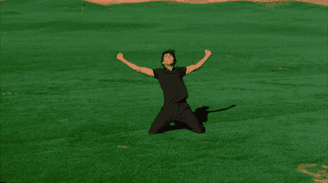 A person in all black clothing celebrates on their knees on a grassy field with arms raised in victory