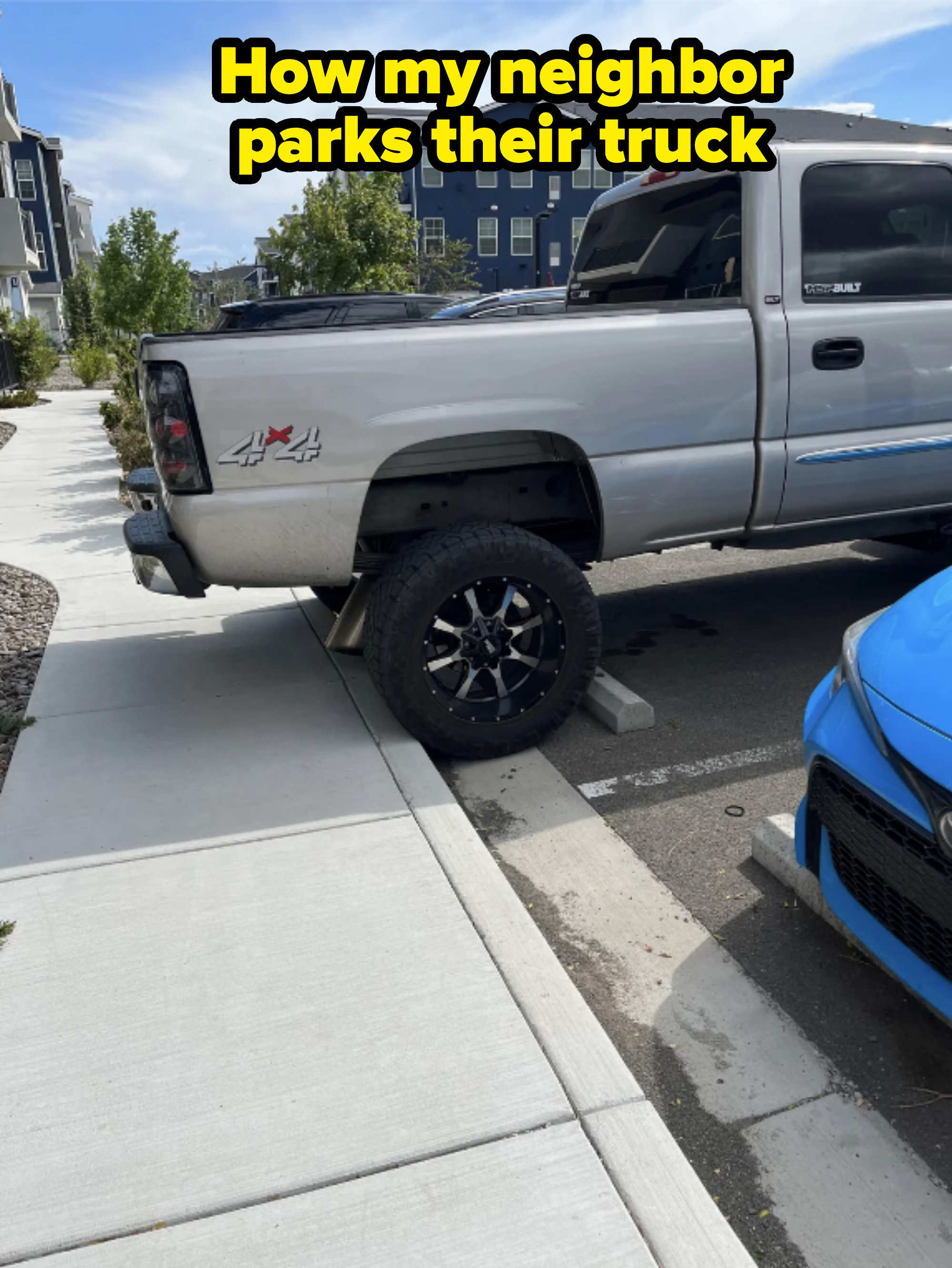 A raised truck with large wheels is parked on a curb, partially on the sidewalk, in a residential neighborhood. A blue car is parked beside it
