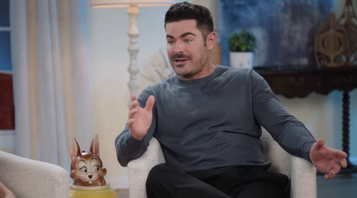 Zac Efron in a casual long-sleeve shirt and pants, sitting and gesturing while speaking during an interview. A Bambi figurine is on the table beside him