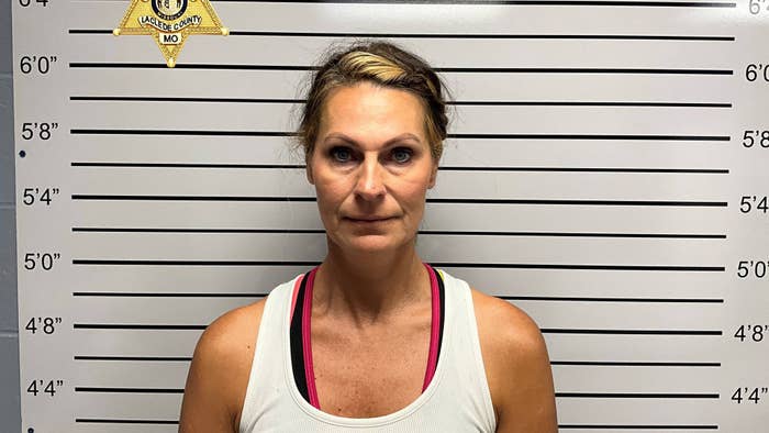 A woman wearing a sleeveless top stands in front of a height chart for a mugshot. The Laclede County, MO sheriff&#x27;s badge is on the upper left