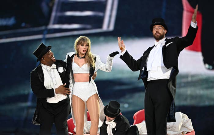 Taylor Swift performs on stage in a white stage outfit with dancers in tuxedos and top hats