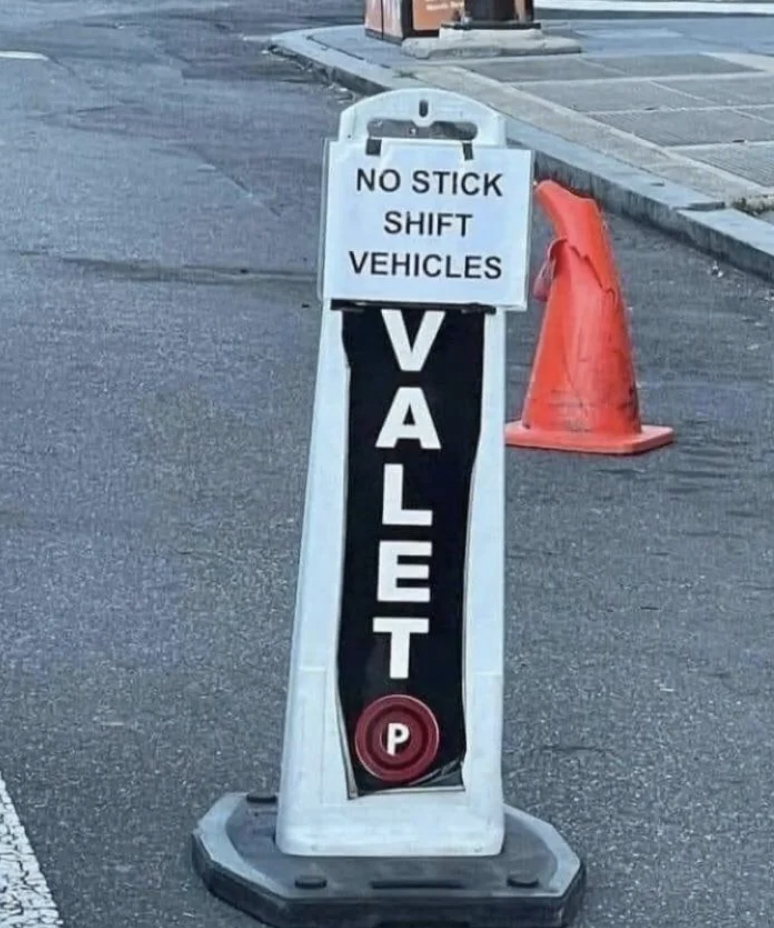 Sign on a street reads &quot;No stick shift vehicles. VALET&quot; with a red parking symbol below. Orange traffic cone on the sidewalk is visible in the background