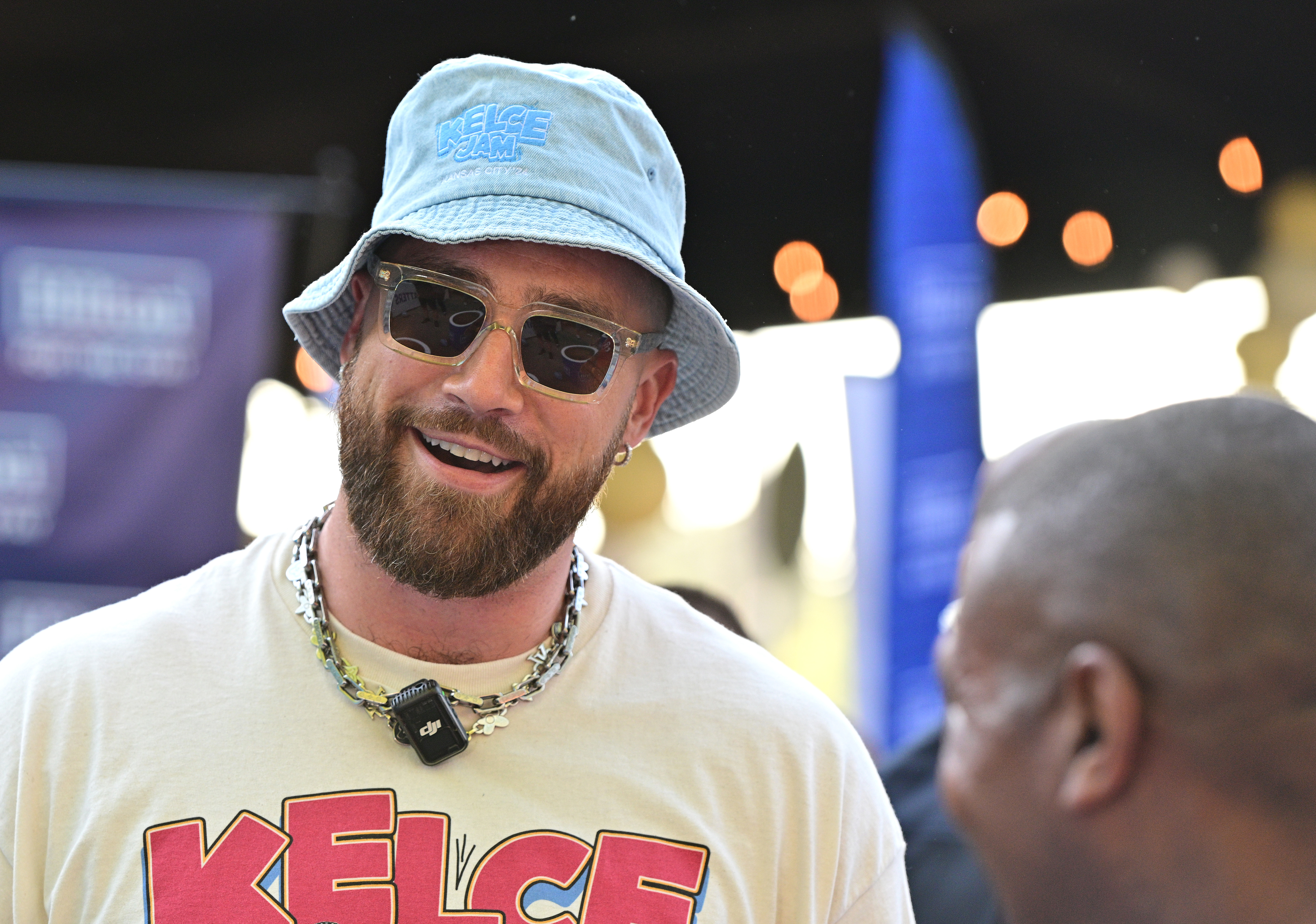 Travis Kelce wearing a blue bucket hat, sunglasses, and a t-shirt with his name "Kelce" on it, smiling at an event