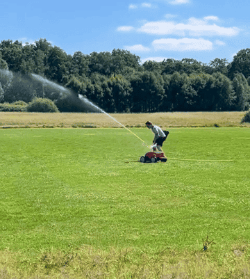 A person mowing a large lawn on a small red mower while being sprayed with water in a park-like setting