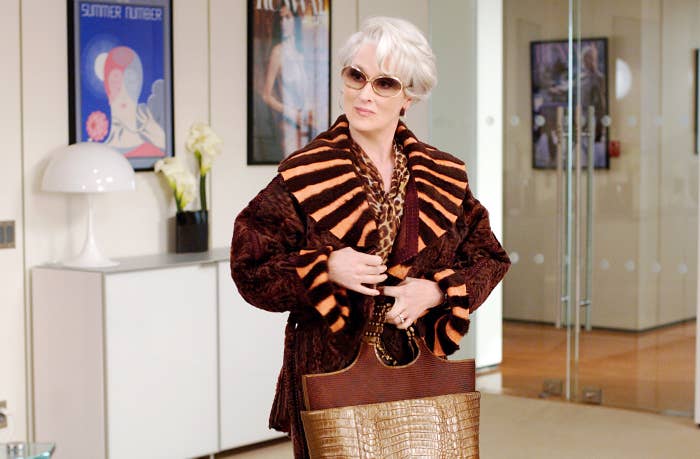 Meryl Streep, wearing large sunglasses, a striped fur coat, and holding a textured handbag, stands confidently in an office setting