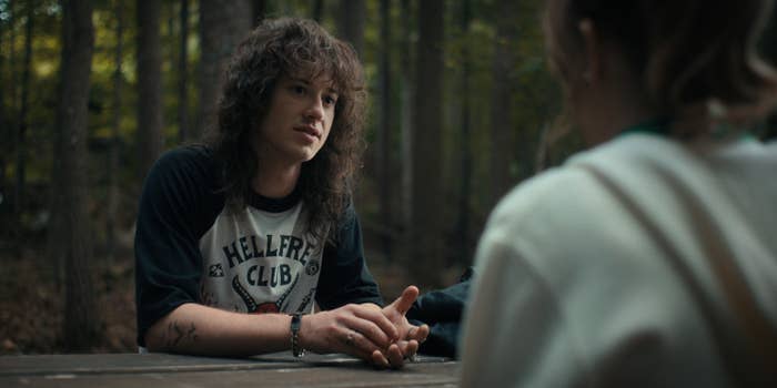 Eddie Munson from "Stranger Things" sits at a wooden table in a forest, talking to an unidentified person