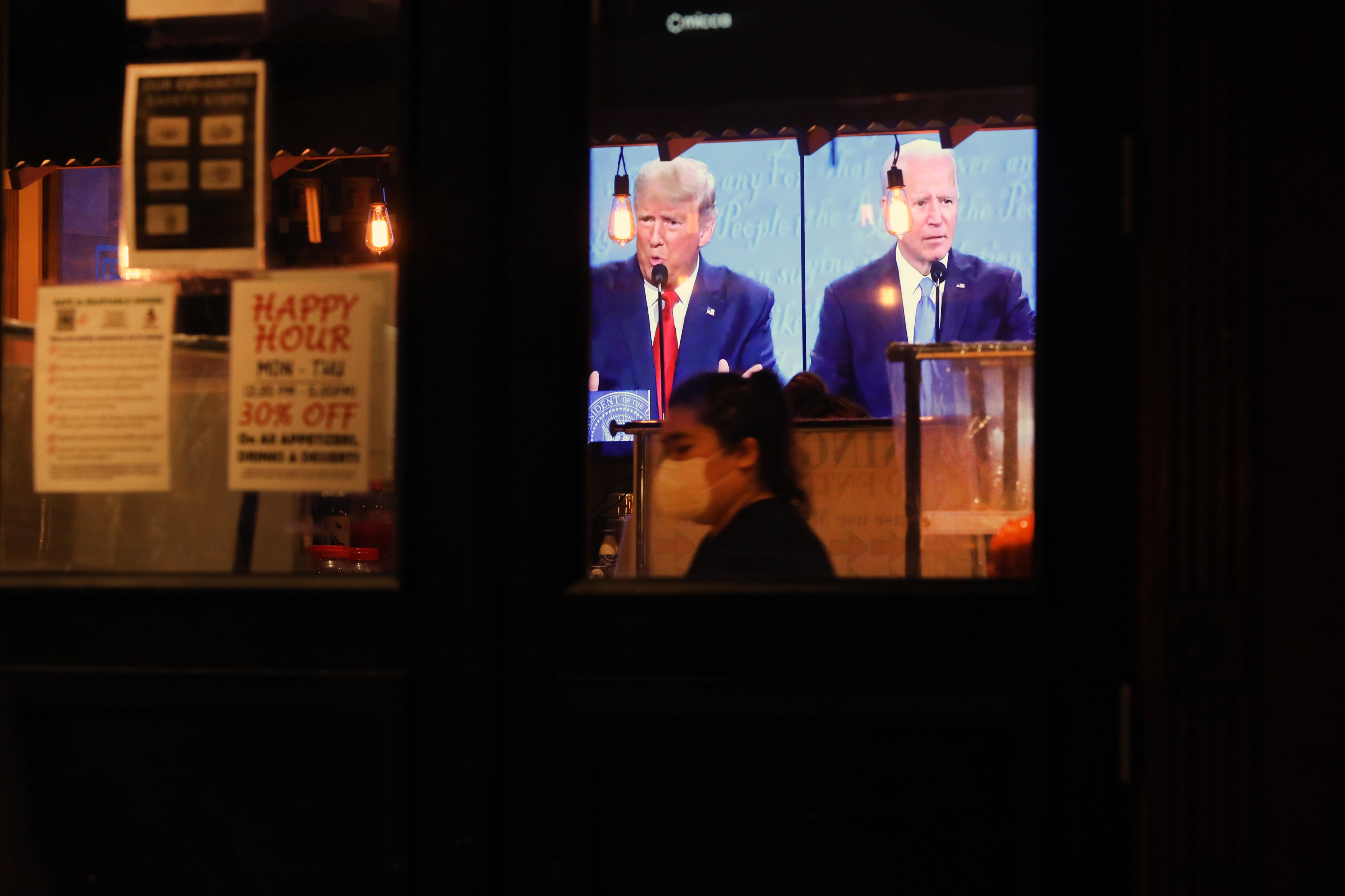 Donald Trump dan Joe Biden muncul di layar TV di dalam restoran yang remang-remang, dengan seseorang bertopeng berdiri di latar depan.