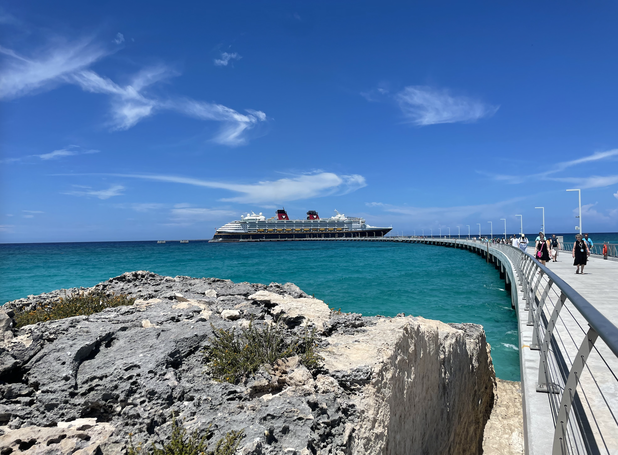 Cruise ship in distance on turquoise ocean, docked at a pier with people walking towards it. Rocky outcrop in foreground under clear sky