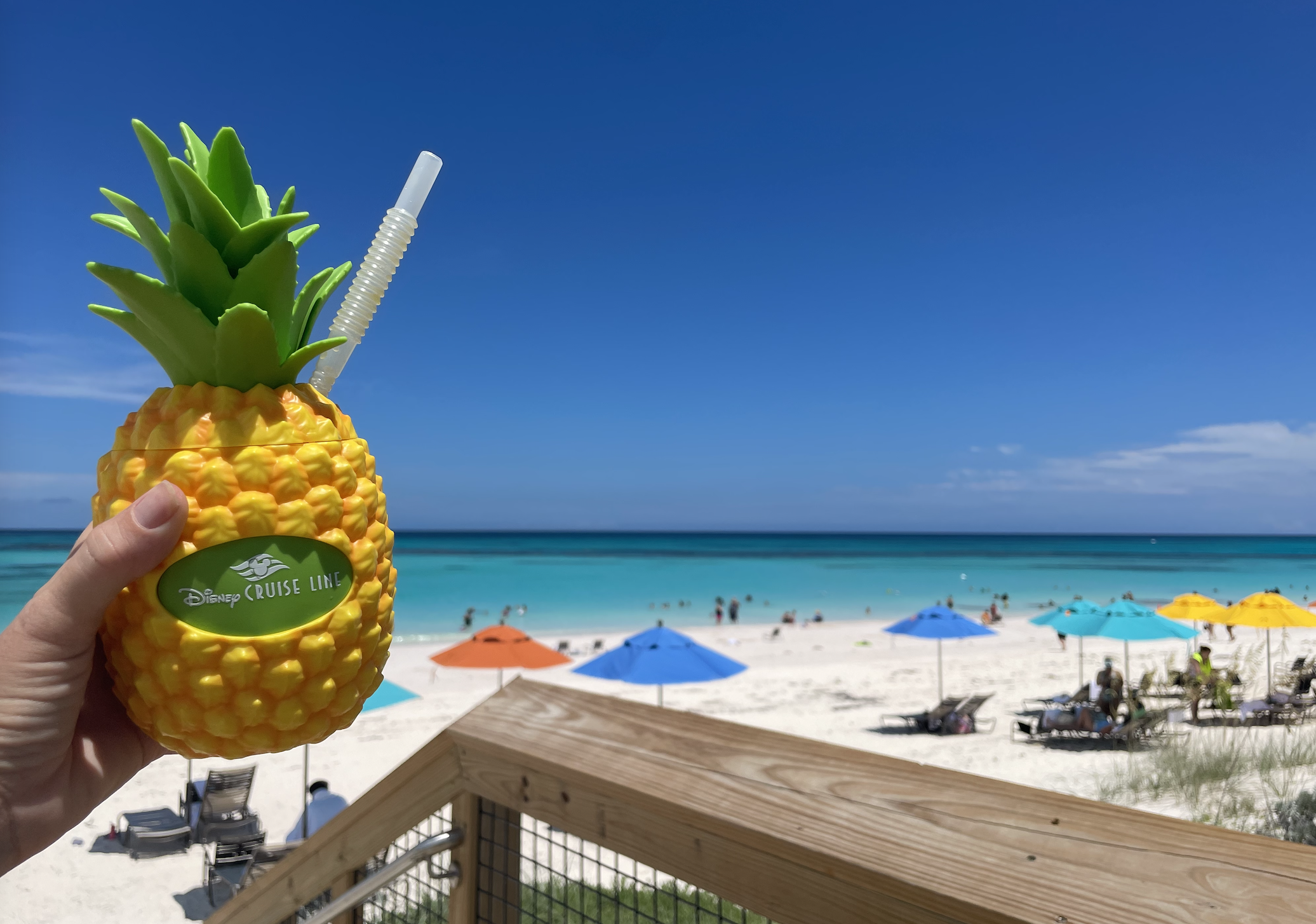 A hand holds a pineapple-shaped Disney Cruise Line cup with a straw on a tropical beach. The beach has colorful umbrellas and people are relaxing by the water
