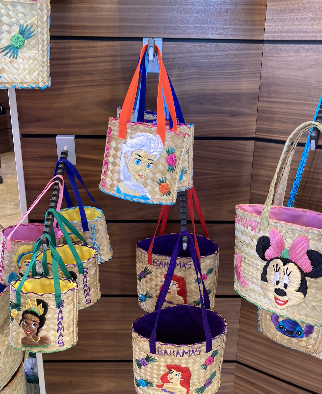 Straw handbags with Disney characters Tiana, Elsa, Ariel, and Minnie Mouse are displayed on wall hooks. Handles are colorful. Bags say &quot;Bahamas.&quot;