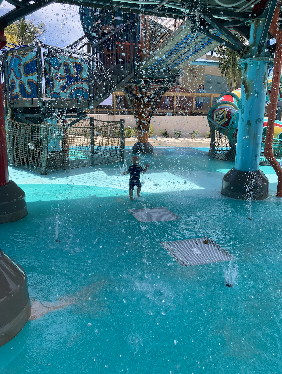 A child is playing under sprinklers in a water playground with colorful, interactive structures around