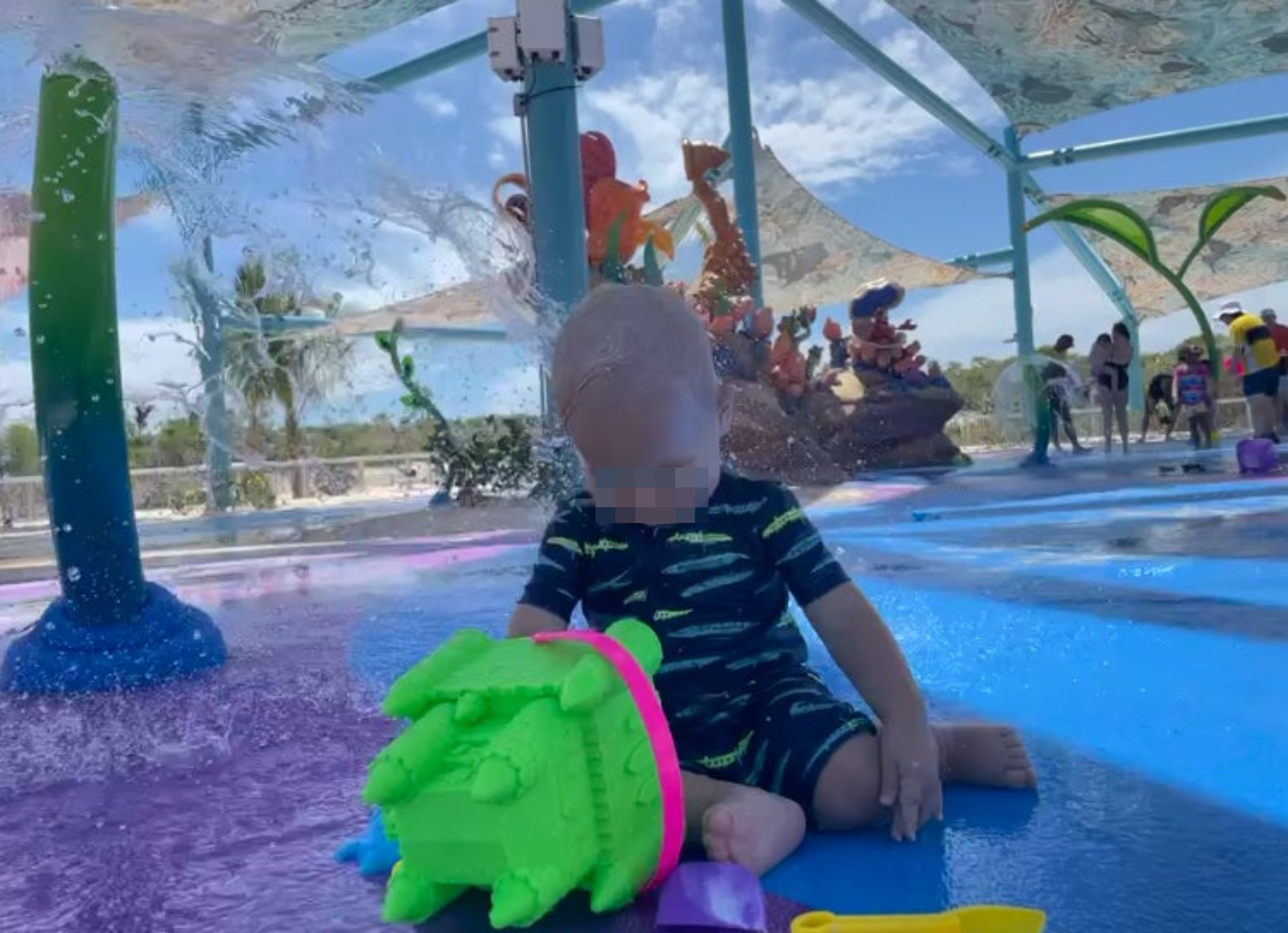 A baby in a splash pad playground, sitting on the ground playing with a green toy, with water splashing on their head from above