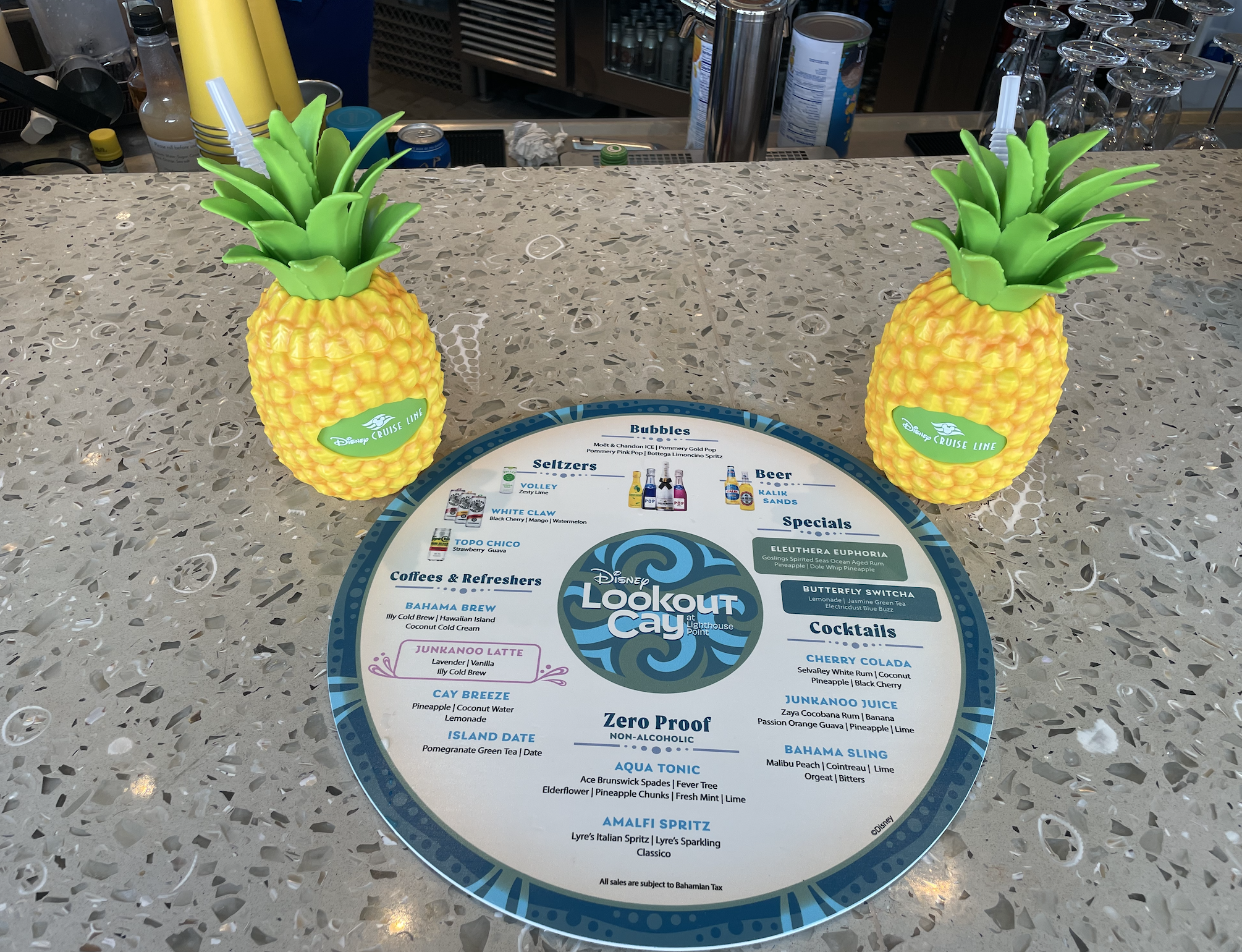 Bar menu with tropical drink options, featuring two pineapple-shaped drink containers on either side of the menu for Disney&#x27;s Lookout Cay