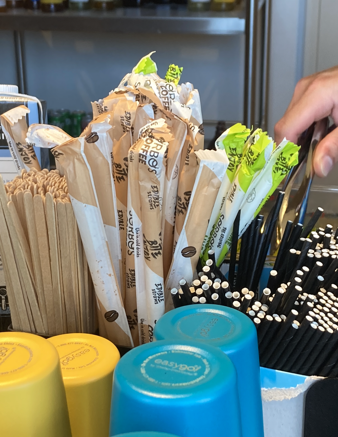 A hand reaches for black straws among a variety of unwrapped and wrapped straws, wooden stir sticks, and upside-down plastic cups in a coffee shop setting