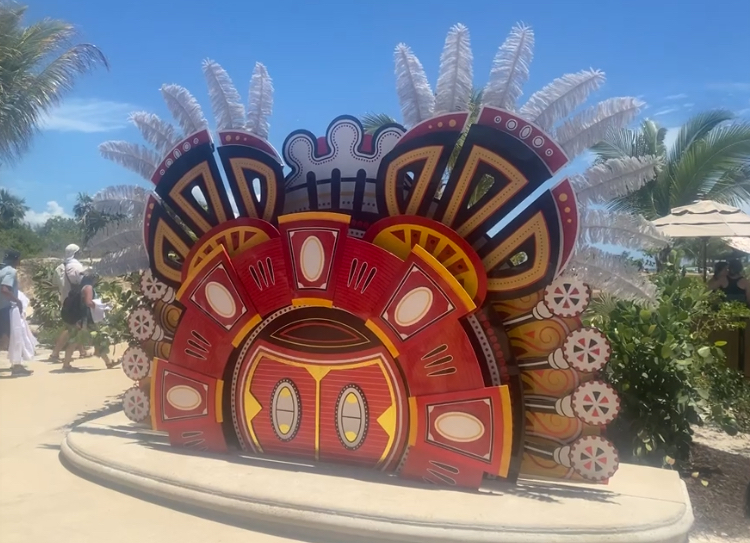 A vibrant, decorative display with intricate geometric patterns and feather-like elements. People walk by in the background under a sunny sky