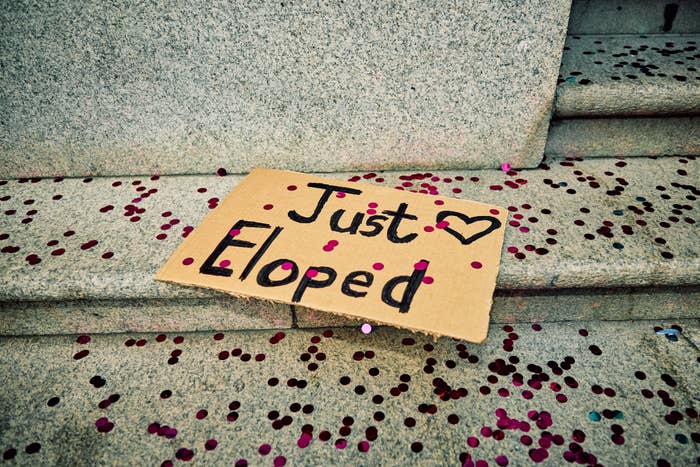 A cardboard sign on steps says "Just Eloped" with a heart drawing, surrounded by scattered confetti