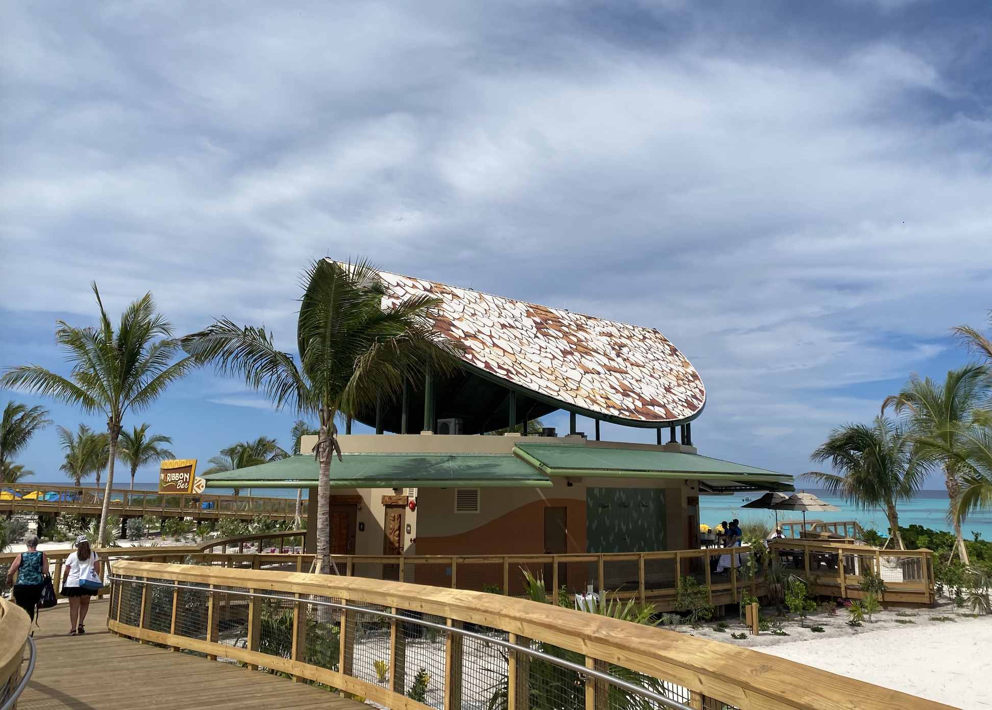 A beachfront hut with a unique curved patterned roof, surrounded by palm trees and a wooden boardwalk with people walking. Ocean visible in the background