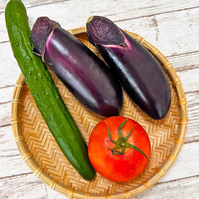 木の表面に置かれた竹の皿の上に、キュウリ、ナス、トマトが並んでいる。
