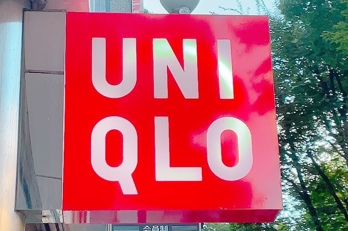 Sign with the text “Uniqlo” on a red background, displayed outdoors next to a building. Trees are visible in the background