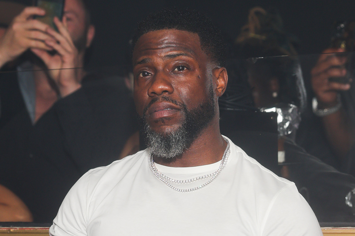 Kevin Hart is at an event wearing a white t-shirt and silver chain necklaces. People in the background are using their phones