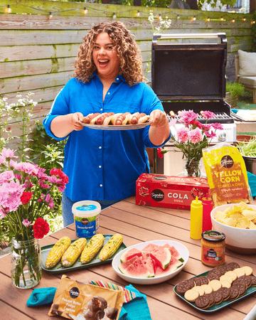 A person setting down a plate of hot dogs on to an outdoor barbecue table set with various foods, drinks, and flowers