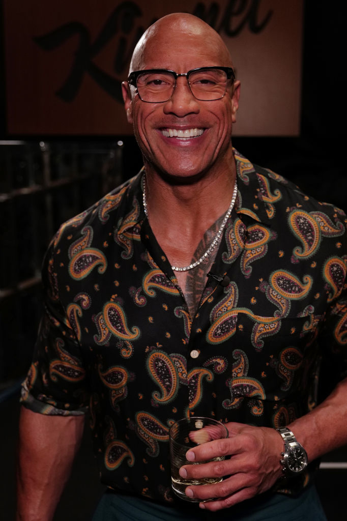 Dwayne Johnson, wearing a paisley-print shirt and glasses, smiles while holding a drink