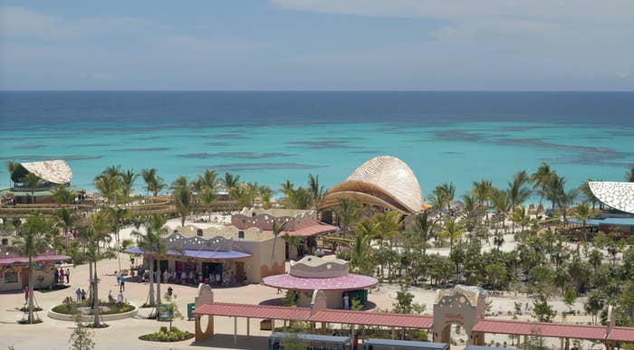 A scenic beach resort with palm trees, a few buildings, and shaded areas for tourists. The clear blue ocean extends into the horizon under a partly cloudy sky