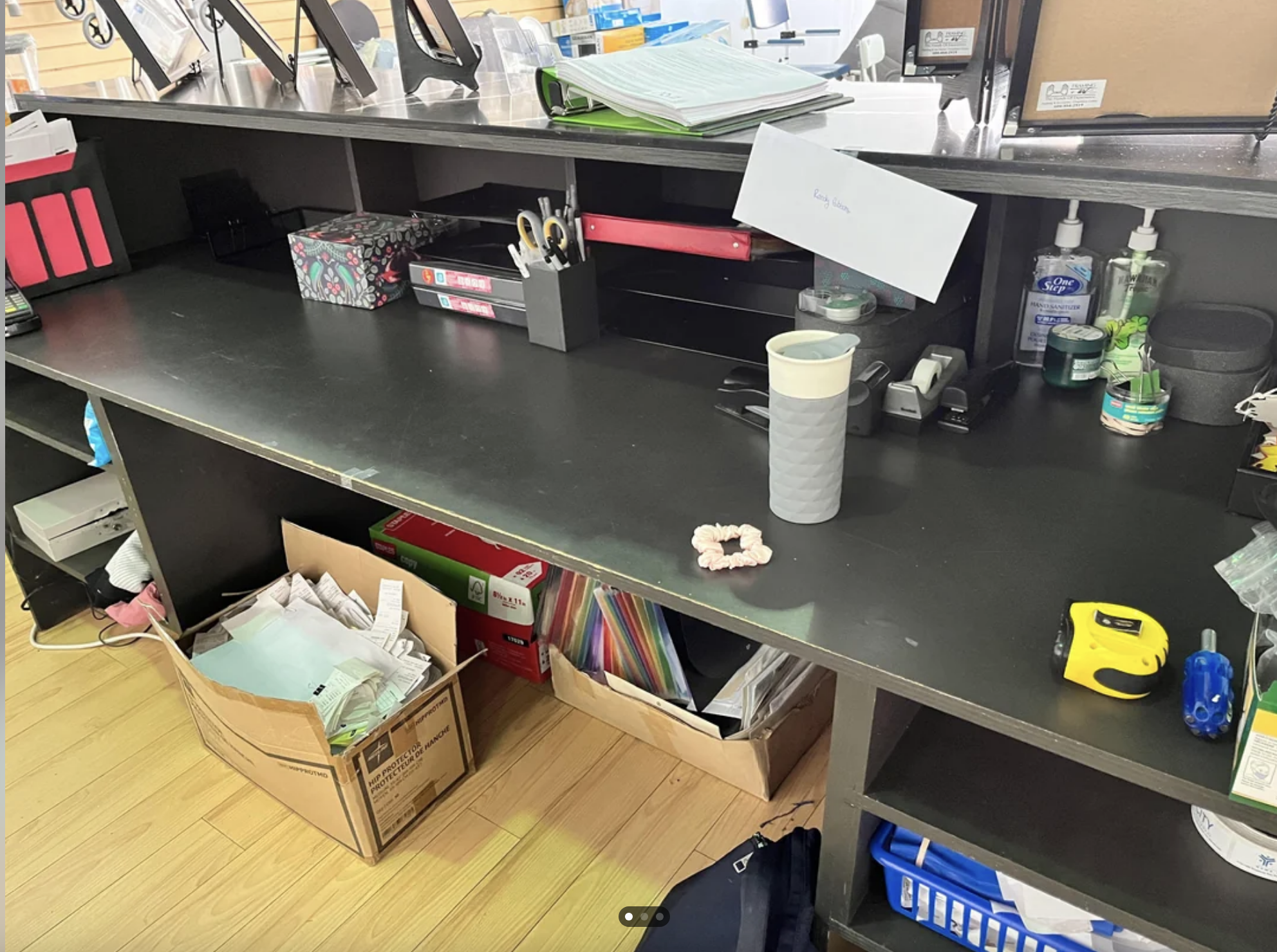 A cluttered office desk with papers, office supplies, and personal items including a coffee cup, tape measure, and a cardboard box filled with documents on the floor