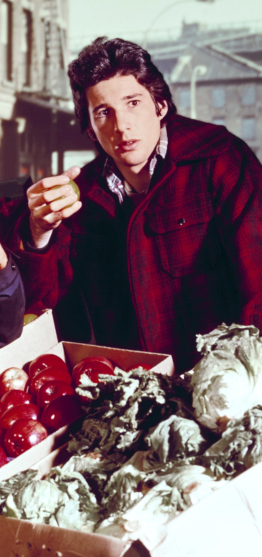 Richard Gere at an outdoor market, interacting with produce. he wears a plaid jacket with a turtleneck