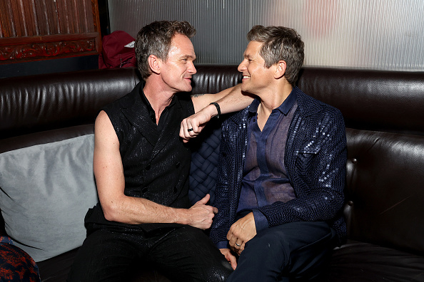 Neil Patrick Harris in a sleeveless black sequin shirt and David Burtka in a blue sequin jacket smile at each other while sitting on a couch