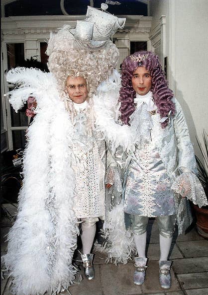 Elton John and David Furnish in elaborate Baroque-style costumes with ornate wigs and feathered cloaks