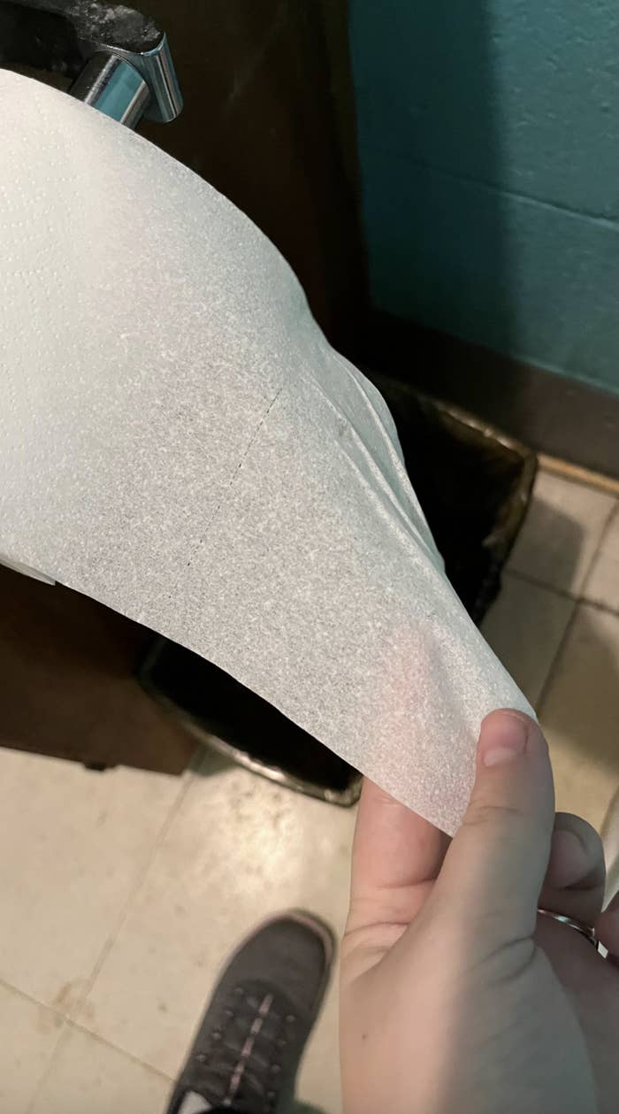 A hand is shown holding a thin, see-through piece of toilet paper, which is unraveling from a dispenser