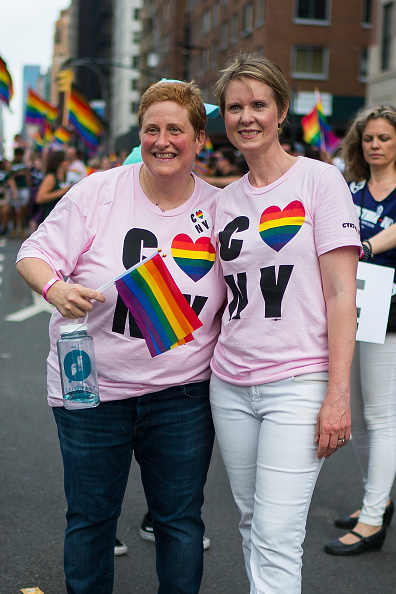 Christine Marinoni and Cynthia Nixon smiling and posing together at a Pride event. They are wearing matching T-shirts with "LOVE NY" and holding a rainbow flag