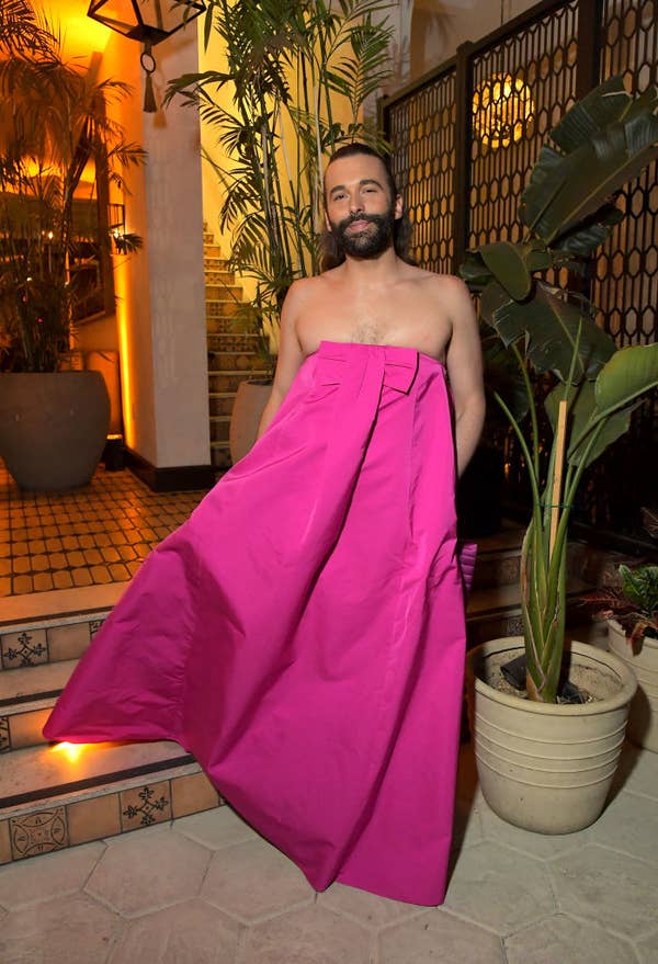 Jonathan Van Ness wears a large fuchsia skirt and is shirtless at a public event. He stands on tiled steps, surrounded by potted plants