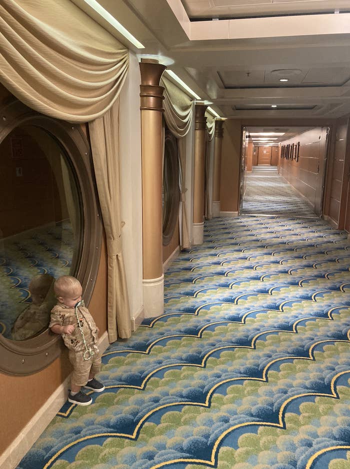 A young child in patterned clothing stands by a large window looking down an ornate hallway with curtains, columns, and a blue and green patterned carpet